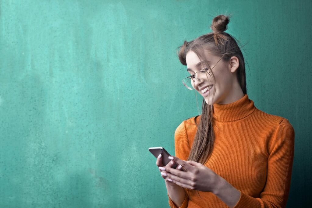 Young woman in orange turtleneck smiling while using smartphone, set against a green wall.